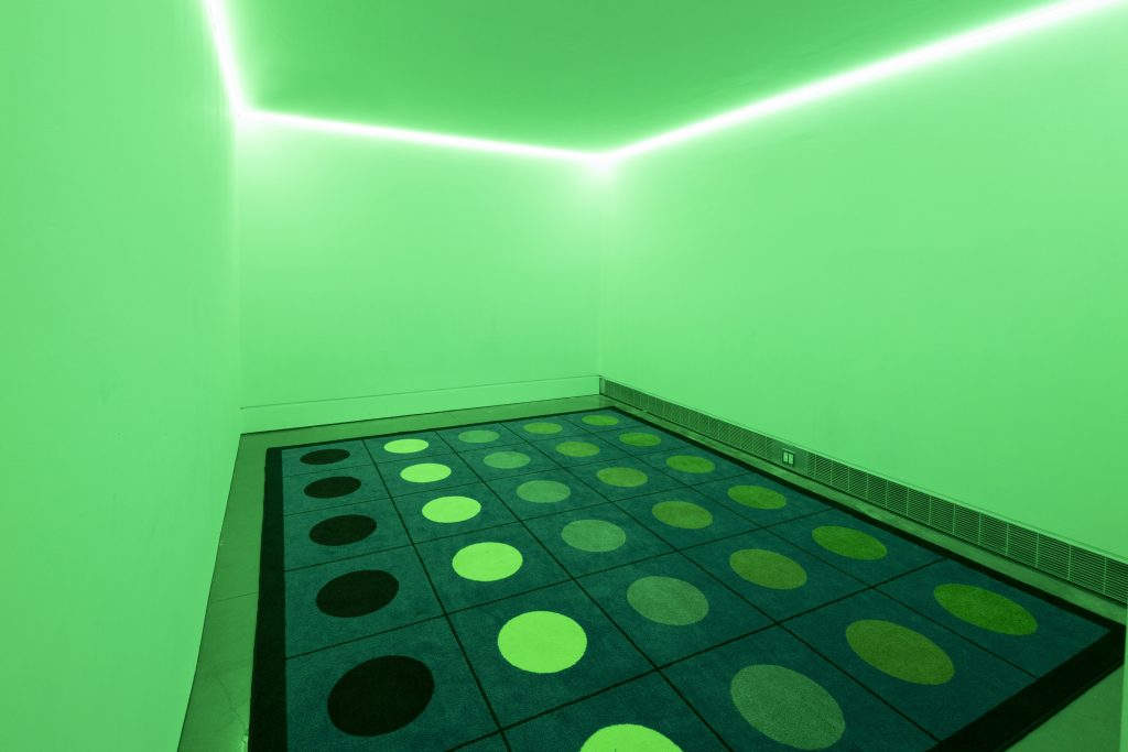 The same room, now lit from above with bright neon green lights, shines on a rug on the floor. The rug has a grid pattern with five rows of six dots in the middle of each grid square. From left to right, each row is now a different color, in different shades of green.