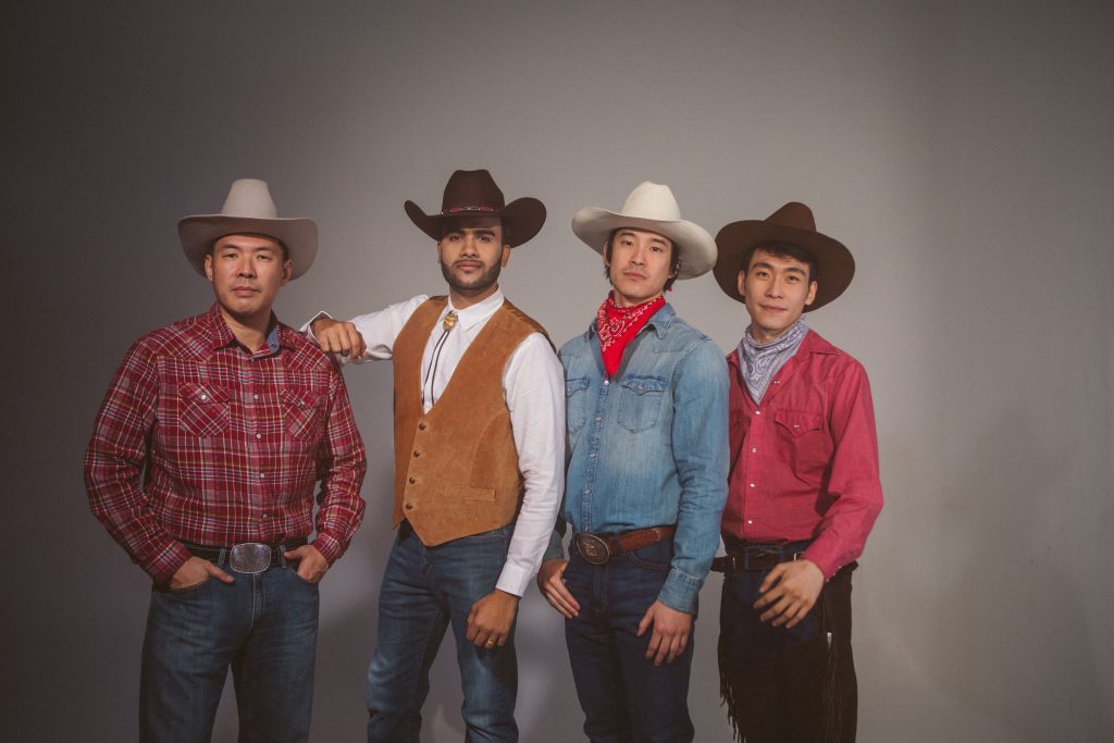 Four men of diverse Asian backgrounds pose shoulder to shoulder. They are dressed in cowboy outfits, with hats, western style shirts, and jeans. 