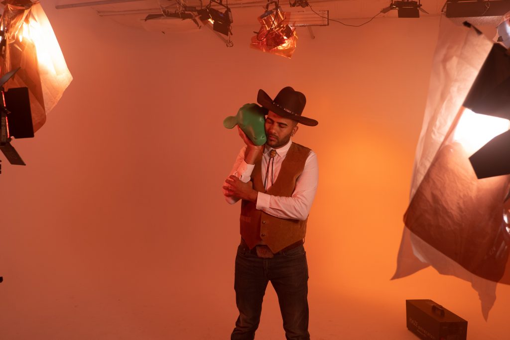 One of the men stands in the center with his eyes closed. He hold a green plastic cow head to the side of his face, tenderly. In the foreground, he is surrounded by stage lights that lend a soft orange glow to the image.