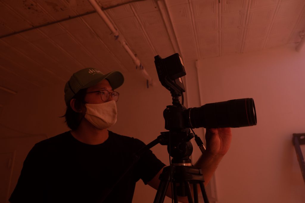 Kenneth Tam, a young Asian male, is pictured dimly lit and in profile, standing in front of a film camera with a long lens. He is wearing a hat, glasses, and a face mask.