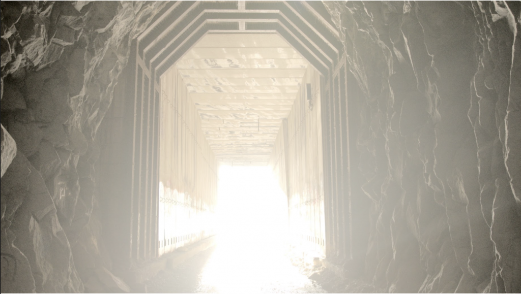 A bright light emanates from the center of a rocky tunnel. A wooden support structure lines its inside. 