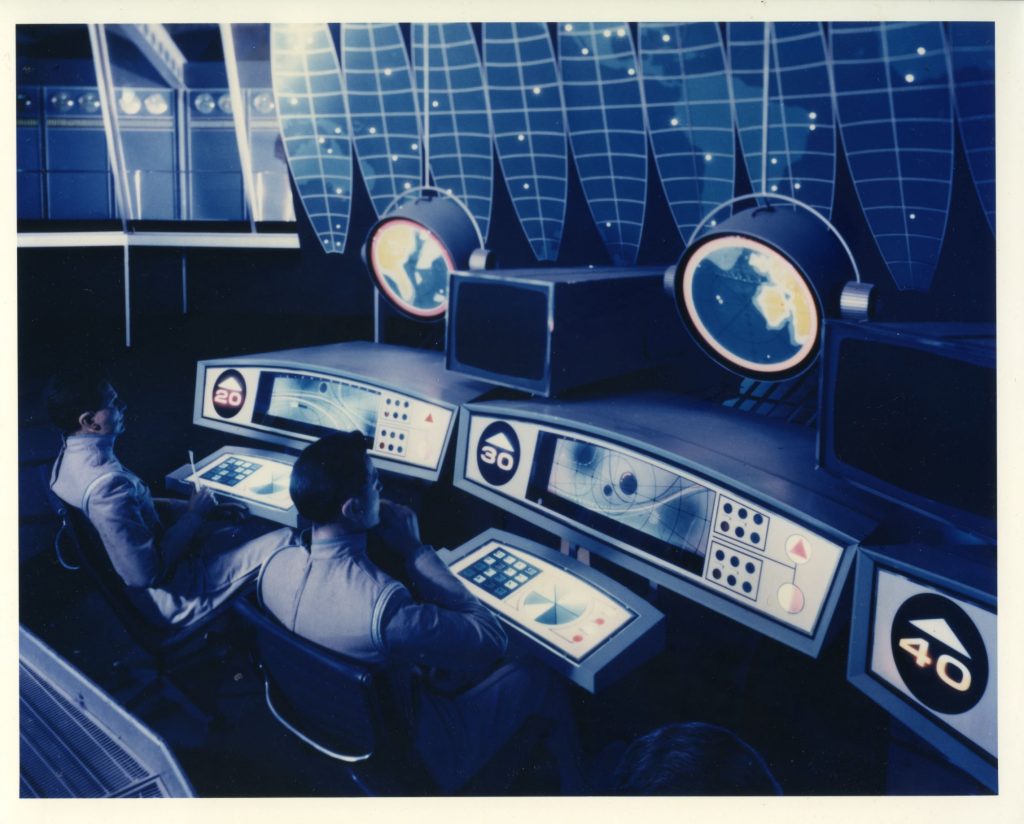 A photograph of the proposed Antarctic Meteorological Station, built and on-display in General Motors’ Pavilion at the 1964-1965 New York World’s Fair as a part of the audiovisual amusement ride, Futurama. Two men sit at a control desk with maps of the world, assorted knobs and buttons, and screens in front of them, assumed to be surveying the environment from beneath the ice, sometime in the future.