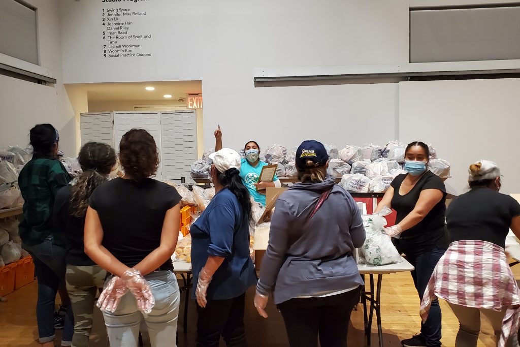 In the foreground, several women stand with their backs to the camera. Many of them wear hats and plastic food handling gloves. The two women who face the camera wear masks and appear focused. The space they stand in is full of tables and bins with plastic bags of food.