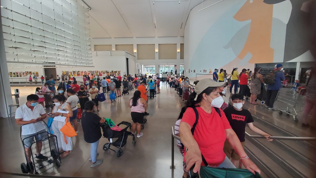 A snaking line of hundreds of individuals and families is pictured in the central atrium of the Queens Museum. Each holds nags, carts, and carriages to receive food within. In the background, artworks by Pedro Felipe Vintmilla Burneo and Ulrike Müller are partially visible.
