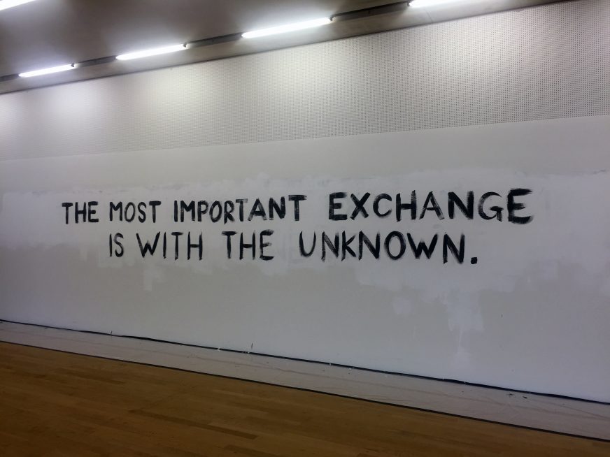 The words “The most important exchange is with the unknown” appear in black capital letters. They are hand drawn on a long horizontal gallery wall. Below is a wooden floor, and above, neon lights.