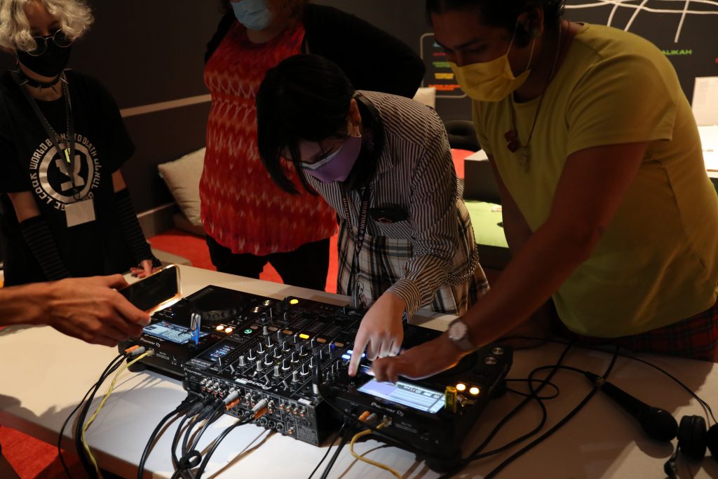 LGBTQ+ teens from Generation Q participate in an introductory DJ Workshop held at the Queens Museum. Two young, stylish people wearing facemasks, point to the illuminated LED screen of DJ turntables. One wears a lime green t-shirt, and the other wears black and white stripes and a checkered skirt. Two other individuals watch in the background, and a hand holding an iPhone as a flashlight illuminates the DJ equipment. 