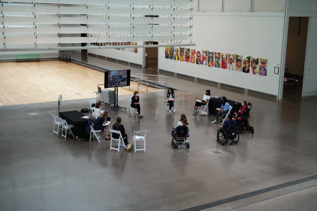 Members of the Access Cohort gathersin the Queens Museum’s Lower Atrium for a Focus Group, dedicated to identifying access challenges posed by the building. Ten individuals wearing face-masks, of varying ages, races, and abilities are seated in a circle, with a large TV monitor on a stand. Three individuals are in wheelchairs and/or motorized scooters. Most people hold clipboards and pens, and look attentively towards one member of the group who is speaking. In the distance, there is a series of colorful banners depicting queer Latinx people, as part of a temporary exhibition. 