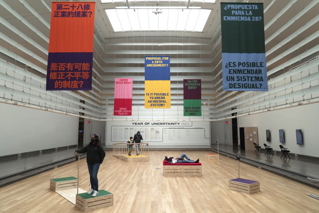 Five colorful banners hang from the ceiling reading “Proposal for a 28th Amendment?” And “Is it possible to amend an unequal system?” in the five most spoken languages in Corona, NY. Below the banners, four visitors stand and lay on the five wooden colorful soapboxes in different arrangements.
