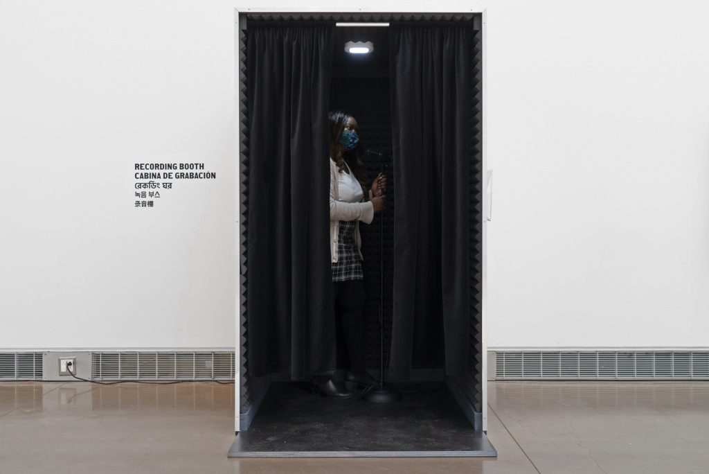 In the center, a visitor is standing in a wooden rectangular recording booth with a black curtain split open. On the wall, to the left of the recording booth reads “Recording Booth”. 