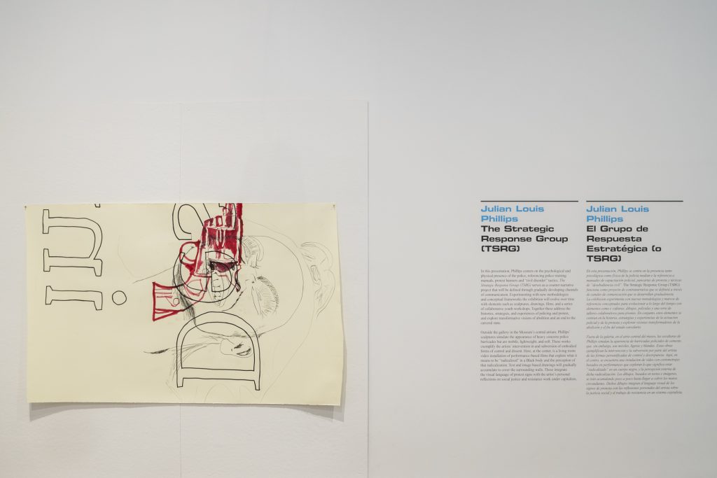 On the left, a drawing by Julian Louis Philips in mixed mediums is pinned to the wall horizontally. On the right, there is the gallery wall text reading the artist's name “Julian Louis Philips”, the exhibition title “ The Strategic Response Group”, and information describing the exhibition and the artist's work. 