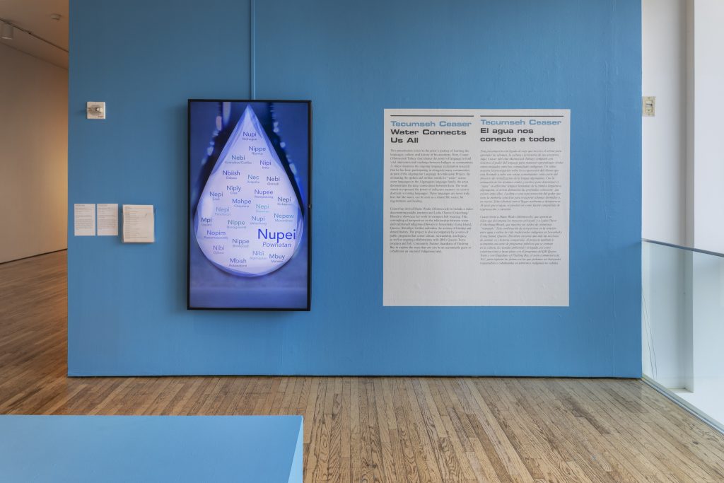 A blue wall has small white wall text on the left. In the middle, a large monitor with a blue water droplet displays ways to say “water” across languages in the Algonquian language family. There is a large white wall text on the right: “Tecumseh Ceaser Water Connects Us All”