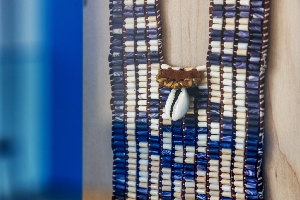 A close up of a woven wampum necklace by Lydia Ann Wallace-Chavez: thick, flat rows of cream shell beads, with blue shell beads woven in a round shape. On the collar is woven brown fabric and a white cowrie shell.