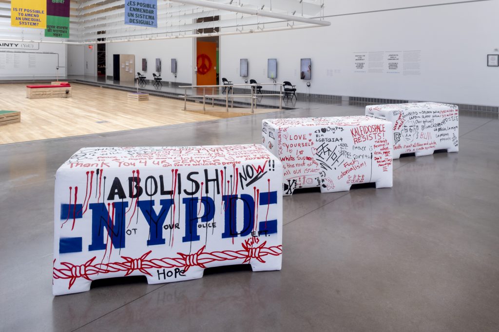 Three white police barricade sculptures with blue N Y P D text and red and black collaborative graffiti on display in the Queens Museum.