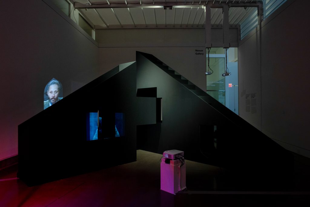 Two huge black triangular sundial structures with stairs contain holes through which videos are projected. A small projector on the floor shines through a square hole in the left triangle, projecting a news anchor’s face onto the opposite wall. Two projectors hang from the ceiling on the right, facing the larger black sculpture.